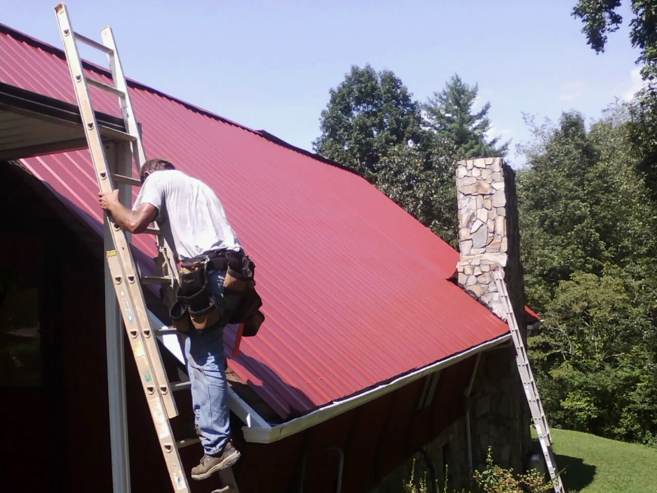 Roofer on a ladder beside a red metal roof and stone chimney — Soffit & Fascia Repair by Roofing Brothers in Incline Village